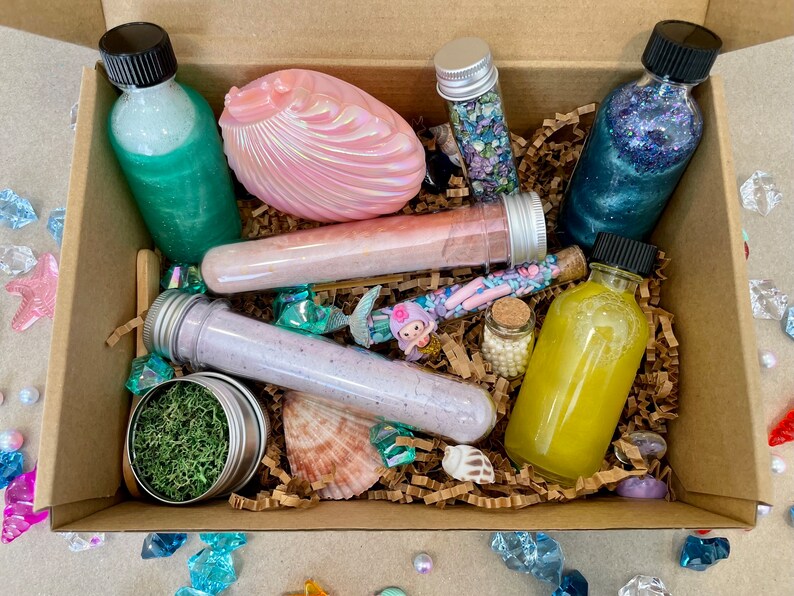 K&ouml;nnte beinhalten: Eine braune Pappbox gef&uuml;llt mit Bade- und K&ouml;rperprodukten, darunter eine rosa Muschel, eine Flasche mit gr&uuml;ner Fl&uuml;ssigkeit, eine Flasche mit blauer Fl&uuml;ssigkeit, eine Flasche mit gelber Fl&uuml;ssigkeit und mehrere R&ouml;hrchen mit Badesalz. Die Box ist umgeben von kleinen Dekorationsartikeln wie Muscheln, Perlen und Kristallen.