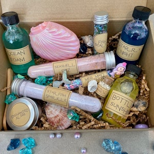 K&ouml;nnte beinhalten: Eine braune Pappbox gef&uuml;llt mit verschiedenen Badeprodukten mit Meerjungfrauen-Thema. Die Box enth&auml;lt eine Muschel, Badesalze, Badebomben und Bade&ouml;l. Die Produkte sind mit Namen wie "Ocean Foam", "Seashell Fizz", "Starfish Sparkle", "Mermaid Magic", "Starry Night" und "Bottle Sunshine" beschriftet.
