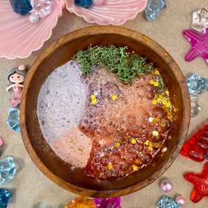 以下が含まれることがあります： ピンク、パープル、オレンジのバスボムの混合物、緑の苔、金色のグリッターが入った木製のボウル。ボウルは、貝殻、ヒトデ、宝石など、さまざまな装飾品で囲まれています。