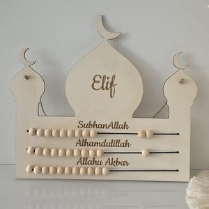 Könnte beinhalten: Ein helles Holzzählwerk in Form einer Moschee mit drei Kuppeln, die von Mondsicheln gekrönt sind. Es zeigt den Namen Elif sowie die Phrasen SubhanAllah, Alhamdulillah und Allahu Akbar und hat drei Reihen verschiebbarer Holzperlen.
