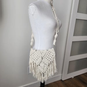 May include: A white crocheted crossbody bag with a fringe detail. The bag is on a white mannequin.