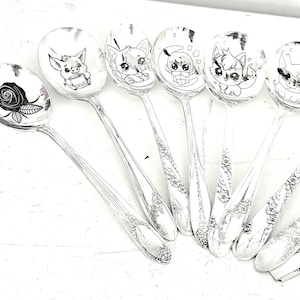 May include: A collection of silver spoons with various hand-drawn illustrations. The spoon bowls feature images of a rose, a pig, and several cartoon cats. The handles have floral patterns.