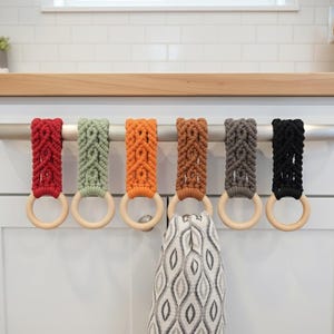 Könnte beinhalten: Sechs Makramee-Handtuchhalter in Rot, Grün, Orange, Braun, Grau und Schwarz. Jeder Halter hat einen Holzring und hängt an einer silbernen Stange. Ein gemustertes Handtuch hängt an einem der Halter. Ideal für die Küche.