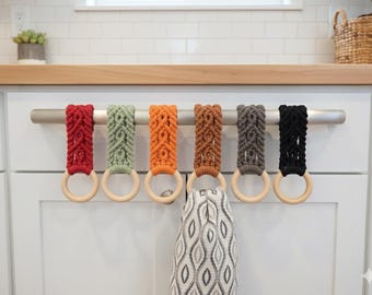 Può includere: Un set di sei portasciugamani in macramè in vari colori: rosso, verde, arancione, marrone, grigio e nero. Ogni supporto ha un anello di legno ed è appeso a una barra d'argento. Un asciugamano fantasia è appeso.