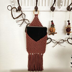 May include: A terracotta-colored macrame wall hanging with a wooden ring at the top. The hanging has a fringe at the bottom and a black book is placed inside the hanging.