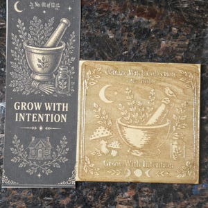 May include: A rectangular black collector card and a square wooden tile from the Cottage Witch Collection. Both feature a mortar and pestle, herbs, a crescent moon, and the text Grow With Intention. The card is labeled No. 01 of 12.