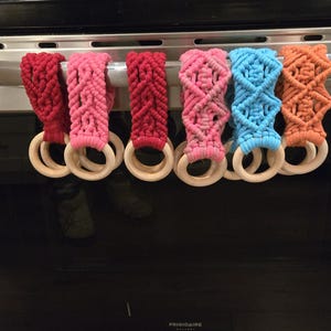 Op de afbeelding: Een verzameling van zes macramé keukendoekhouders in verschillende kleuren, waaronder rood, roze, blauw en oranje. Elke houder heeft een houten ring en hangt aan een roestvrijstalen ovenhandvat.