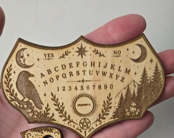 Könnte beinhalten: Ein helles Holz-Spirit-Board mit lasergravierten Details, darunter das Alphabet, die Zahlen 1-9 und 0, die Wörter YES, NO und GOODBYE, ein Rabe, Mondsicheln, Sterne, ein Bergwald und ein Pentagramm, begleitet von einer passenden Planchette.