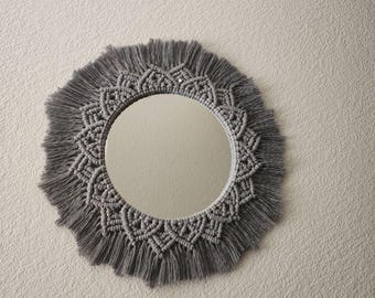 Puede incluir: Un espejo de macramé gris con un espejo circular y un borde de flecos.