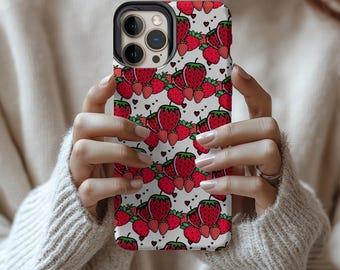 May include: A phone case featuring a repeating pattern of red strawberries with green leaves and small black hearts on a white background. The case is held in front of a person wearing a cream-colored sweater.