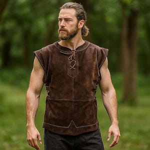 Pode incluir: Um colete de camurça castanho com decote com atacadores e ombreiras. O colete tem um bolso e é usado sobre uma camisa preta. O fundo é uma floresta desfocada.