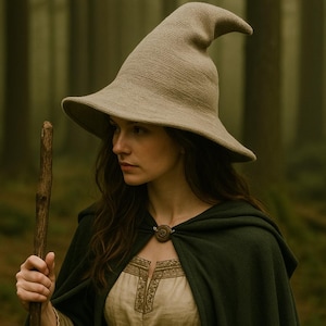 May include: A woman wearing a beige witch hat with a pointed crown and wide brim, a dark green cloak, and a light-coloured dress with gold embroidery. She is holding a wooden staff in a forest setting.