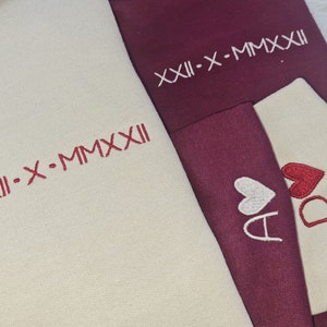 May include: Two cream-colored hoodies with red embroidery. One hoodie has the text "XIII-X-MMXXII" and the other has two hearts with the letters "A" and "D" inside.