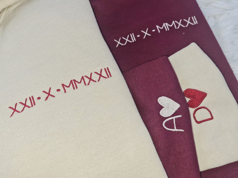 May include: Two cream-colored hoodies with red embroidery. One hoodie has the text "XIII-X-MMXXII" and the other has two hearts with the letters "A" and "D" inside.