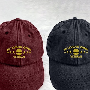 May include: Two baseball caps with an embroidered skull and crossbones design. The caps are black and burgundy with gold embroidery. The text on the caps reads "Malevelon Creek" and "Veteran".