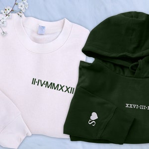 May include: A set of two sweatshirts, one white and one dark green. The white sweatshirt has the Roman numeral date "II-IV-MMXXII" embroidered on the front and a heart with the letter "R" on the sleeve. The dark green sweatshirt has a hood and the Roman numeral date "XXVI-III-MMXXII" embroidered on the front and a heart with the letter "S" on the sleeve.