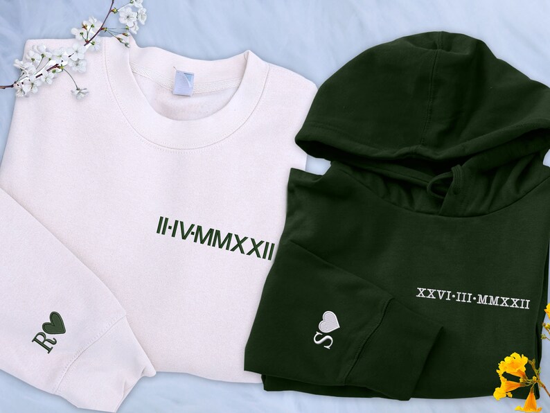 May include: A set of two sweatshirts, one white and one dark green. The white sweatshirt has the Roman numeral date "II-IV-MMXXII" embroidered on the front and a heart with the letter "R" on the sleeve. The dark green sweatshirt has a hood and the Roman numeral date "XXVI-III-MMXXII" embroidered on the front and a heart with the letter "S" on the sleeve.
