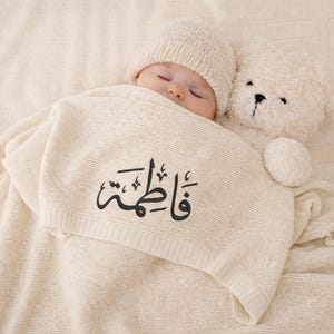 Könnte beinhalten: Ein schlafendes Baby mit einer cremefarbenen Strickmütze, eingehüllt in eine cremefarbene Decke. Auf der Decke steht der Name "Fatima" in schwarzer Schrift. Ein cremefarbener Teddybär liegt neben dem Baby.