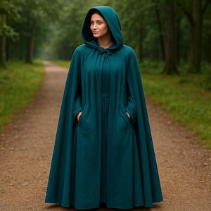 May include: A teal hooded cloak with a long, flowing design. The cloak features a hood, a button closure at the neck, and pockets. The person is standing on a path in a forest.