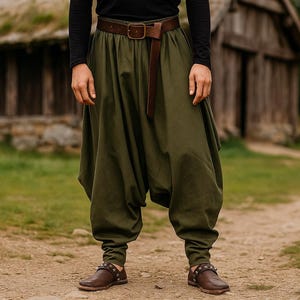 Könnte beinhalten: Olivgrüne Haremshose mit braunem Ledergürtel und passenden Schuhen. Die Hose hat ein lockeres, fließendes Design, das an den Knöcheln gerafft ist. Der Gürtel hat eine rechteckige Schnalle. Im Hintergrund befindet sich eine rustikale Holzstruktur.