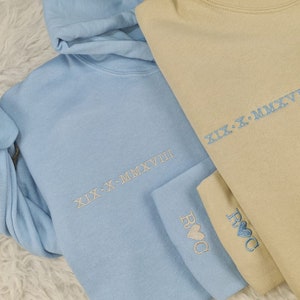 May include: Two hooded sweatshirts, one light blue and one beige, with the same embroidered text "XIX-X-MMXVIII" and the text "ROC" on the sleeves.