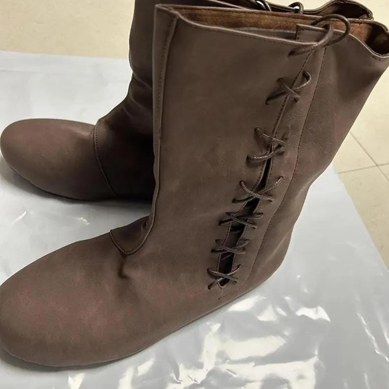 May include: A pair of brown leather boots with a mid-calf height. The boots have a rounded toe and a side lace-up design. The leather has a matte finish, and the boots appear to be unworn.