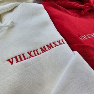 May include: Two hooded sweatshirts, one white and one red, with red embroidered Roman numeral dates. The white sweatshirt has "VIII.XII.MMXXI" and the red sweatshirt has "VIII.XI.MMXXI".