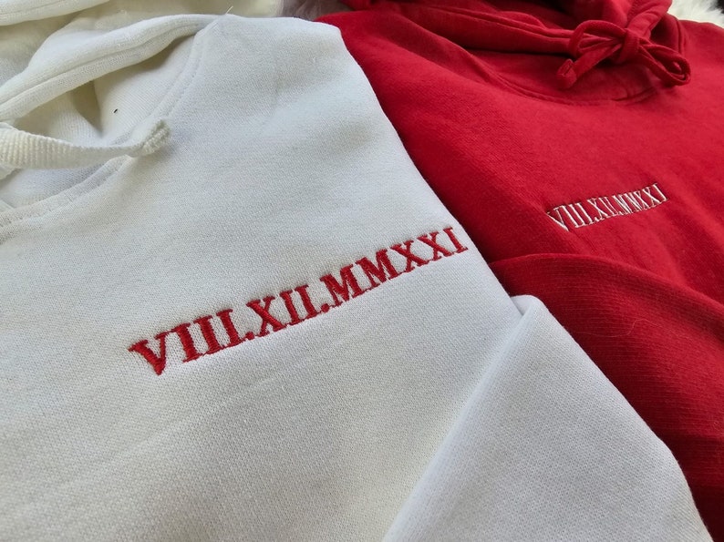 May include: Two hooded sweatshirts, one white and one red, with red embroidered Roman numeral dates. The white sweatshirt has "VIII.XII.MMXXI" and the red sweatshirt has "VIII.XI.MMXXI".