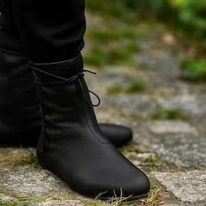 May include: Black leather ankle boots with a lace-up closure. The boots have a rounded toe and are worn with black pants. The boots are on a stone surface with green foliage in the background.