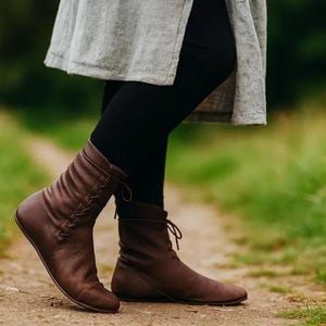 May include: Brown leather ankle boots with a lace-up design. The boots have a low heel and are worn with black leggings and a gray tunic. The boots are on a dirt path with green grass on either side.