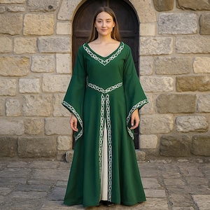 May include: A long, emerald green dress with wide sleeves and a V-neck. The dress has white embroidered trim on the neckline, waist, and sleeves. A person is wearing the dress, standing in front of a stone wall.