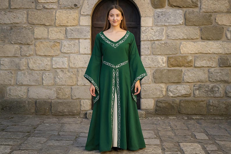 May include: A long, emerald green dress with wide sleeves and a V-neck. The dress has white embroidered trim on the neckline, waist, and sleeves. A person is wearing the dress, standing in front of a stone wall.