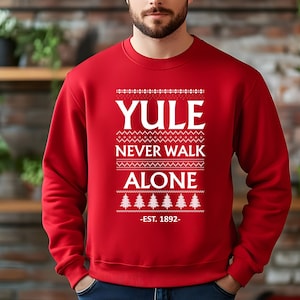 May include: Red sweatshirt with the text "YULE NEVER WALK ALONE - EST. 1892" in white. The design includes decorative patterns and Christmas trees. A festive holiday-themed apparel item.