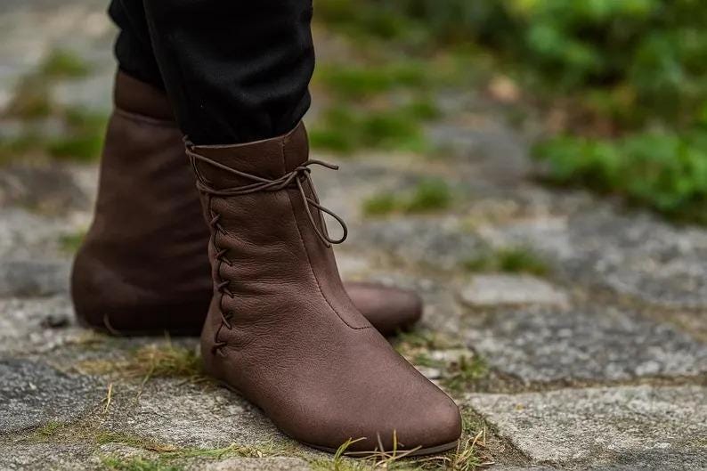 May include: Brown leather ankle boots with a lace-up design. The boots have a rounded toe and a flat sole. The image shows the boots from the ankle down, with the boots on a stone surface.