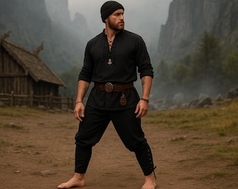 May include: A person wearing a black long-sleeved shirt with a lace-up neckline, black pants, and a brown belt with a decorative buckle. The person is also wearing a black beanie and standing barefoot on a dirt path.