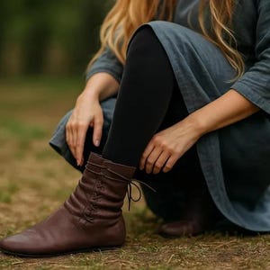 May include: Brown leather ankle boots with a lace-up design. The boots have a low heel and are worn with black leggings and a long-sleeved blue-grey dress. The image is taken outdoors on a grassy surface.