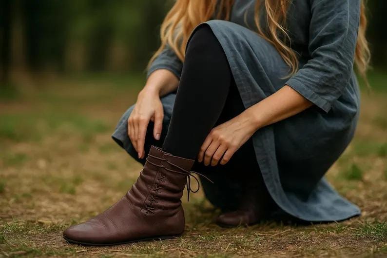 May include: Brown leather ankle boots with a lace-up design. The boots have a low heel and are worn with black leggings and a long-sleeved blue-grey dress. The image is taken outdoors on a grassy surface.