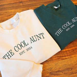Może przedstawiać: Dwie bluzy z napisem "The Cool Aunt Est. 2024" w zielonym kolorze. Jedna bluza jest kremowa, a druga ciemnozielona.