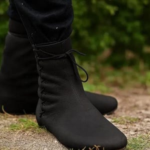May include: A pair of black leather ankle boots. The boots have a rounded toe, a lace-up closure, and stitching details. The boots are worn outdoors on a dirt path with green foliage in the background.
