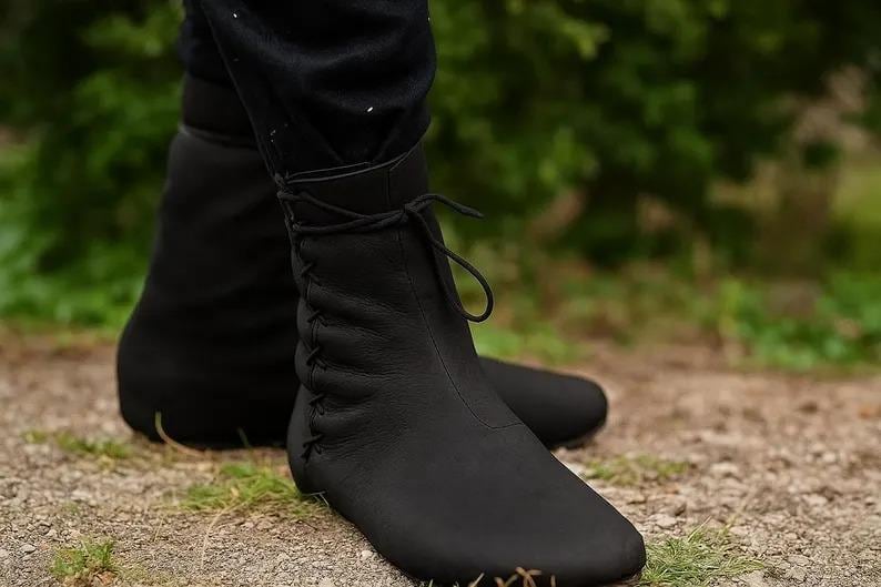 May include: A pair of black leather ankle boots. The boots have a rounded toe, a lace-up closure, and stitching details. The boots are worn outdoors on a dirt path with green foliage in the background.