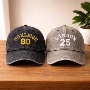 May include: Two baseball caps with curved brims sit side-by-side. One is black with "HUXLEIGH 80" embroidered in yellow. The other is gray with "LANDON 25" embroidered in white. Both caps are made of a worn, distressed fabric.