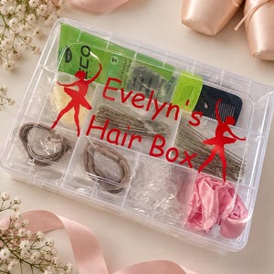 May include: A clear plastic rectangular organizer box labeled "Evelyn's Hair Box" in red text with two red ballerina silhouettes. The box contains hair accessories including bobby pins, hair ties, a small comb, and a green tube.