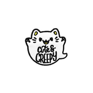 May include: Embroidered patch featuring a white ghost-shaped cat with black outlines. The cat has yellow ears and a smiling face. The text "cute & creepy" is embroidered in black on the ghost's body. The patch is designed for clothing or accessories.