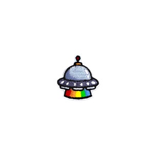 May include: Embroidered patch depicting a cartoon UFO. The UFO has a light blue top, white trim, and a rainbow-coloured base. A small red antenna is visible. The patch is designed to be ironed or sewn onto fabric.