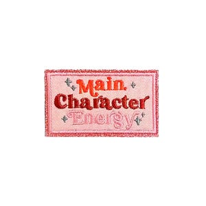 Puede incluir: Parche rectangular rosa con las palabras "Main Character Energy" bordadas en hilo rojo y rosa. El parche tiene un borde rosa brillante y pequeños detalles de estrellas.