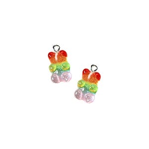 May include: A pair of rainbow gummy bear charm earrings. The earrings are made of silver and feature a gummy bear charm in the shape of a bear. The gummy bear charm is made of clear plastic and has a rainbow colour scheme. The earrings are ideal for adding a touch of fun to any outfit.