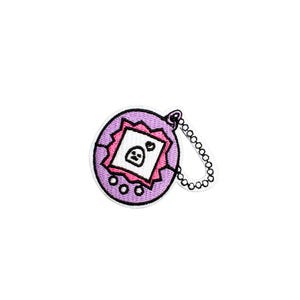 May include: A purple embroidered patch in the shape of a retro handheld digital pet game. The patch features a white screen with a sad face, a pink border, and a silver chain. The patch is a nostalgic accessory.