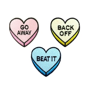May include: Three heart-shaped embroidered patches with black borders. One is pink with "GO AWAY" in black text. Another is yellow with "BACK OFF" in black text. The third is blue with "BEAT IT" in black text.