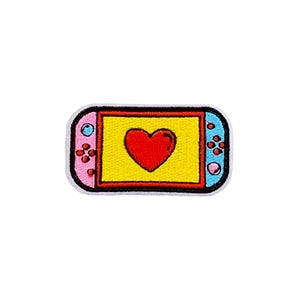 May include: Embroidered patch featuring a cartoon-style video game console. The console has a yellow screen with a red heart, pink and blue sides, and black buttons. The patch is outlined in white.