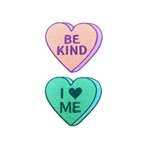 May include: Two heart-shaped patches with embroidered messages. The top patch is pink with purple lettering that says "BE KIND". The bottom patch is green with dark green lettering that says "I ♥ ME".
